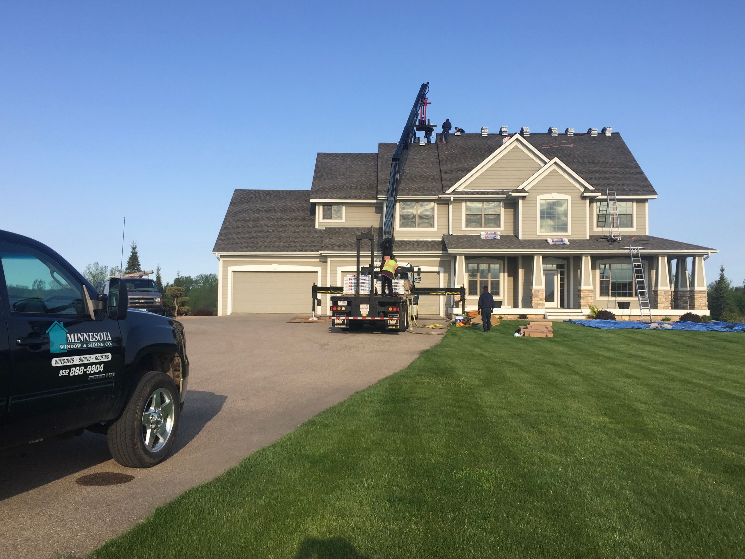 Boom truck lifting shingle bundles to the roof of a two-story home, company pickup at the curb